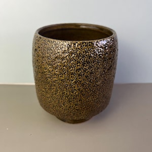 May include: A textured, bronze-colored ceramic pot with a wide opening. The surface has a detailed, organic pattern. The pot has a rounded shape and a slightly flared base. The interior is a darker shade of brown.