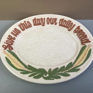 May include: Oval ceramic serving platter with the words "Give us this day our daily bread" in brown lettering. The rim features green leaves and yellow and green corn stalks. The plate has a speckled, off-white background.