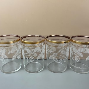May include: Four clear drinking glasses with gold rims and a white grape leaf design. The glasses are cylindrical and appear to be vintage, suitable for serving beverages. The glasses are arranged in a row on a light blue surface.