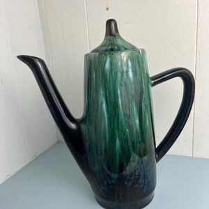 May include: A ceramic coffee pot featuring a glossy glaze with a drip pattern in shades of deep forest green, emerald, and black. The vessel has a tall, cylindrical body, a pointed lid, a long curved spout, and a sturdy handle on the side.