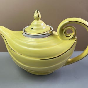May include: A yellow ceramic teapot with a gold-trimmed lid and handle. The teapot has a unique, curved handle and a decorative finial on the lid. The teapot is a light yellow color with gold accents.