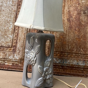 May include: A table lamp with a glossy gray ceramic base featuring embossed floral patterns and two vertical cutouts. The lamp is topped with a rectangular off-white fabric shade and includes a white electrical cord with a plug at the end.