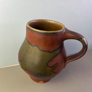 May include: Handmade ceramic mug with a unique design. The mug features a brown handle and rim, with a body adorned with olive green and reddish-brown organic shapes. The mug is a perfect addition to any kitchen or coffee collection.