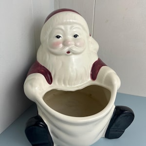May include: A ceramic Santa Claus planter with a cream-colored body, burgundy accents, and black boots. The planter has a large opening for holding items. The Santa figure has rosy cheeks and a white beard. A festive holiday decoration.