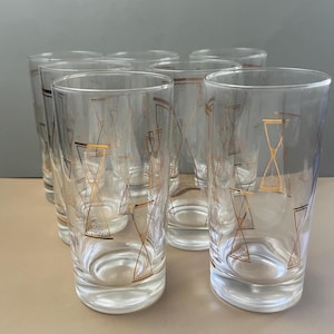 May include: Set of clear highball glasses with gold hourglass and geometric designs. The glasses have a simple, elegant shape, perfect for serving cocktails or other beverages. The gold accents add a touch of vintage charm to the glassware.