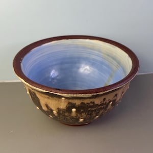 May include: Handmade ceramic bowl with a brown rim and exterior, and a light blue interior. The bowl has a textured surface with small, white, raised dots. The base of the bowl is red.