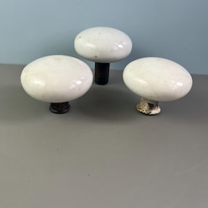 May include: Three white ceramic cabinet knobs with a glossy finish. The knobs have a rounded, oval shape and are mounted on dark metal stems. The knobs are arranged against a light blue and gray background.