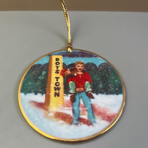 May include: A round Christmas ornament with a gold trim and a gold string. The ornament features a winter scene with a person in a red jacket and blue pants, holding another person. The words "BOYS TOWN" are on a yellow sign.