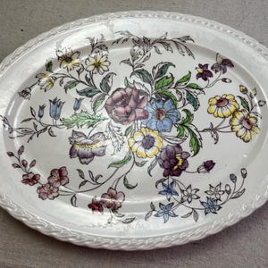 May include: Oval serving platter with a white background and a floral design. The flowers are in shades of purple, yellow, blue, and pink, with green leaves. The rim has a textured, rope-like design.