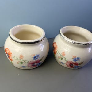 May include: Two small, round, cream-colored ceramic vases with a floral design featuring red, blue, and pink flowers. Each vase has a silver rim. The vases are displayed on a gray surface against a blue background.