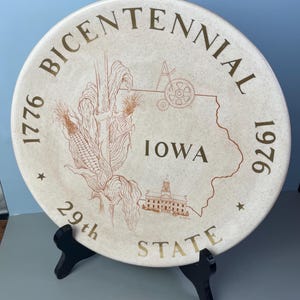 May include: A cream-colored plate commemorating the Bicentennial of 1776-1976. The plate features an outline of Iowa, corn, a building, and the text "29th State". The plate is displayed on a black stand.