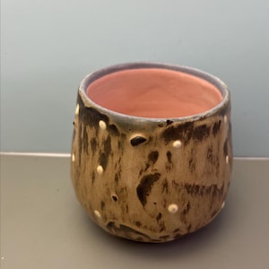 May include: A handmade ceramic cup with a rounded shape. The exterior is tan with dark brown and white dot accents. The rim is a darker color, and the interior is a light pink.