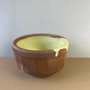 May include: A handmade ceramic bowl with a textured, brown exterior and a smooth, light yellow interior. The rim has a darker brown accent, and the yellow glaze drips down the side. The bowl has a rustic, artisanal aesthetic.