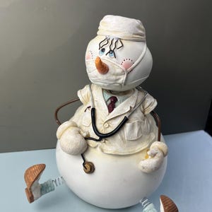 May include: A snowman figurine dressed as a medical professional, wearing a white surgical cap, a face mask, and a white lab coat over a shirt with a red tie. The figure features a carrot nose, a black stethoscope, and spring-attached feet.