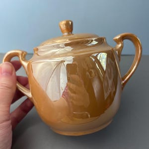 May include: A peach-colored sugar bowl with a glossy, iridescent finish. The bowl features a rounded body, two curved side handles, and a matching lid with a small knob handle on top. The surface reflects light with a metallic sheen.