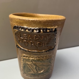 May include: A tan ceramic tumbler with a textured surface. The upper section features a dark brown rectangular plaque with the embossed text "LEADVILLE NORDIC". Below this, a second rectangular plaque displays an embossed skier in motion.