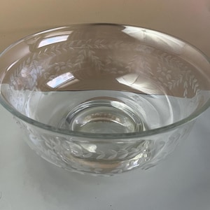 May include: Clear glass bowl with etched leaf and vine pattern. The bowl has a wide, shallow design with a rounded base and a decorative etched pattern around the rim and the base. The bowl is transparent, allowing light to pass through.