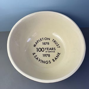 May include: Cream-colored ceramic bowl with a dark brown rim. The bowl features the text "MAPLETON TRUST & SAVINGS BANK" with "1878" and "1978" and "100 YEARS OF SERVICE" in black lettering. The bowl is round and has a smooth surface.