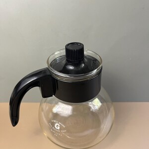 May include: A clear glass coffee carafe with a black plastic handle, lid, and collar. The glass body features the white printed text GEMCO BRAND, a G logo, Heat Resistant Glass, Made in USA, and the number 06 on the front surface.