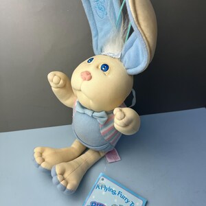 May include: A plush rabbit toy with cream-colored skin, blue eyes, and a pink nose. It wears blue and pink striped overalls with a light blue bowtie. The rabbit has long ears, one upright and one folded, with a signature on the inner ear. A tag reads A Flying, Furry Tale. Bunnybees. An Xavier Roberts.