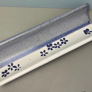 May include: A ceramic, rectangular tray with a curved design. The tray features a light blue and white color scheme, with a dark blue stripe and floral pattern. The tray has a hole on one end.