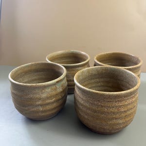 May include: A set of four small, round ceramic cups with a textured, earthy brown and tan speckled glaze. Each cup features a series of horizontal ridges along the exterior body, a wide circular opening, and a smooth, rounded rim design.
