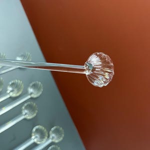 May include: Clear glass stirring spoons with a decorative, scalloped design on the spoon head. The spoons are arranged on a gray surface with a reddish-brown background. The spoons are transparent and reflect light.