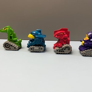 May include: Four colorful toy vehicles with tank treads. The toys are green, blue, red, and purple, each with a unique design. The toys are on a white surface.