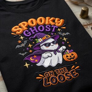Può includere: Una t-shirt nera con un ricamo raffigurante un fantasma bianco con un cappello da strega viola e occhiali da sole. Il fantasma tiene in mano un secchiello a forma di zucca. Il testo recita SPOOKY GHOST ON THE LOOSE, circondato da pipistrelli e stelle.