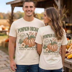 Può includere: Una t-shirt a maniche corte color crema con una grafica di una zucca e il testo PUMPKIN ORCHARD Est. 2021, ORCHARD PICKED, LOCALLY HARVESTED e PUMPKIN PIE APPLE CIDER HAYRIDE stampato in inchiostro verde scuro e marrone sul davanti.