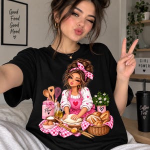 Può includere: Una t-shirt nera con un'illustrazione grafica di una persona con fascia e grembiule rosa che impasta. Il design include un barattolo di farina rosa, un cestino di pane, utensili da cucina e piccoli cuori rosa su un panno a quadretti.