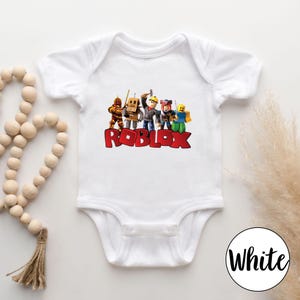 May include: White baby bodysuit featuring a colorful graphic of Roblox characters and the word "ROBLOX" in red block letters. The bodysuit has short sleeves and a snap closure. A white circle with the word "White" is in the bottom right corner.