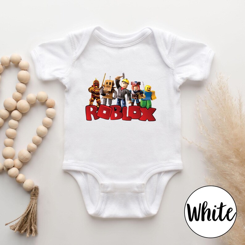 May include: White baby bodysuit featuring a colorful graphic of Roblox characters and the word "ROBLOX" in red block letters. The bodysuit has short sleeves and a snap closure. A white circle with the word "White" is in the bottom right corner.