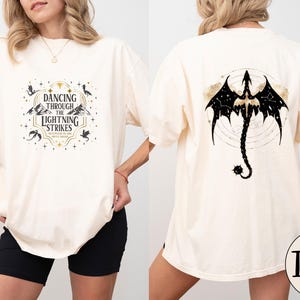 May include: Ivory-colored t-shirt with a front graphic that reads "Dancing Through The Lightning Strikes." The back features a black dragon design with a constellation pattern. The shirt is a comfortable, oversized fit.