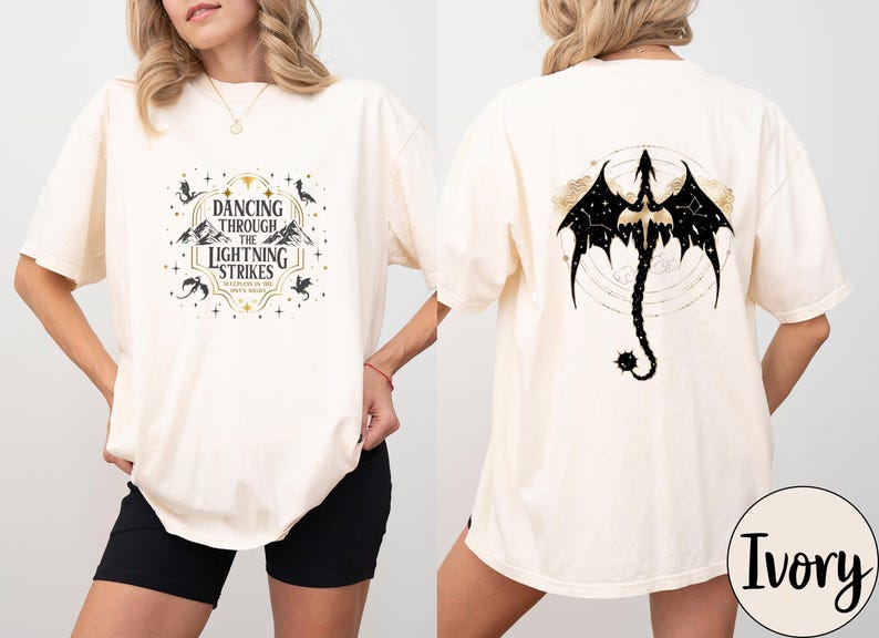May include: Ivory-colored t-shirt with a front graphic that reads "Dancing Through The Lightning Strikes." The back features a black dragon design with a constellation pattern. The shirt is a comfortable, oversized fit.