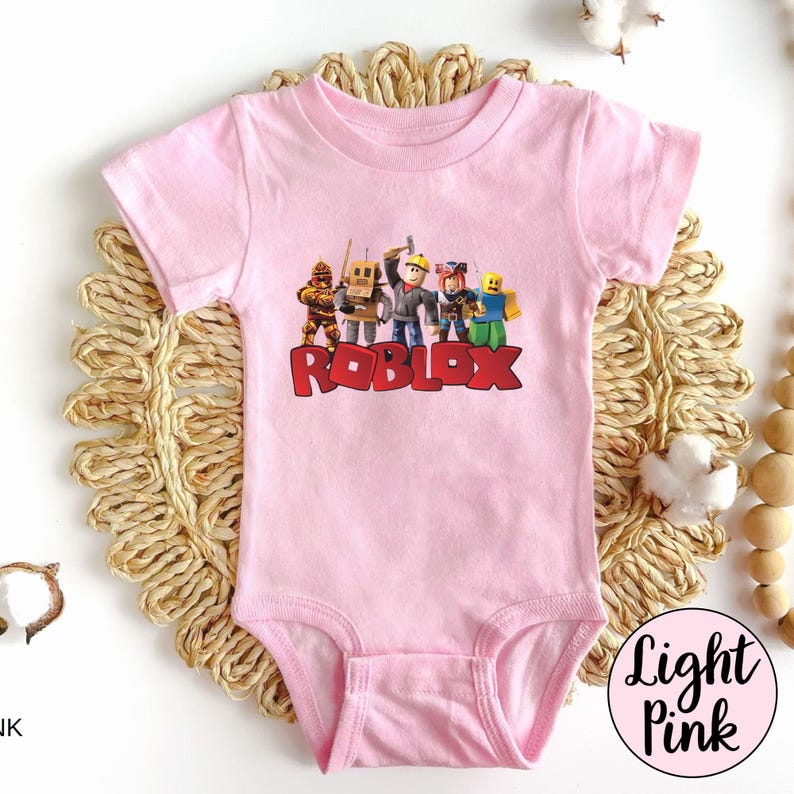 May include: Light pink baby bodysuit featuring a colorful graphic of Roblox characters and the Roblox logo in red. The short-sleeved onesie is made of soft material.
