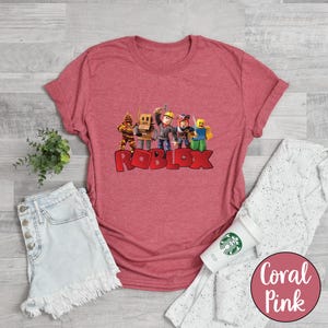 May include: Coral pink t-shirt featuring a graphic of Roblox characters and the Roblox logo in red. The shirt is displayed with denim shorts, a white sweater, and a coffee cup.
