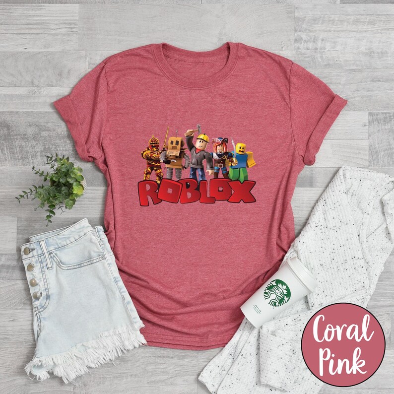 May include: Coral pink t-shirt featuring a graphic of Roblox characters and the Roblox logo in red. The shirt is displayed with denim shorts, a white sweater, and a coffee cup.