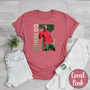 Op de afbeelding: Koraalroze t-shirt met een afbeelding van een voetballer in een rood shirt, groene shorts en een handtekening. Het nummer 7 is zichtbaar. Het woord "RONALDO" staat verticaal in groen en wit gedrukt.