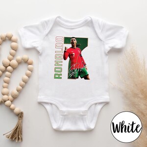 Op de afbeelding: Witte babybody met een afbeelding van een voetballer in een rood shirt met nummer 7. Het woord "RONALDO" is verticaal in groen en rood gedrukt. Inclusief een handtekening.