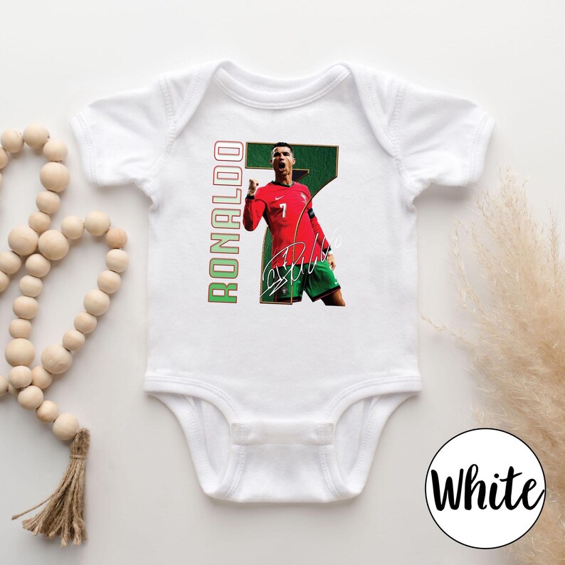 Op de afbeelding: Witte babybody met een afbeelding van een voetballer in een rood shirt met nummer 7. Het woord "RONALDO" is verticaal in groen en rood gedrukt. Inclusief een handtekening.