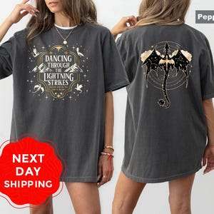 May include: Pepper-colored t-shirt with a front graphic that reads "Dancing Through The Lightning Strikes" and a back graphic of a dragon. The shirt has a relaxed fit and short sleeves. The text is in white and gold.