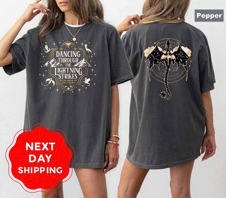 May include: Pepper-colored t-shirt with a front graphic that reads "Dancing Through The Lightning Strikes" and a back graphic of a dragon. The shirt has a relaxed fit and short sleeves. The text is in white and gold.