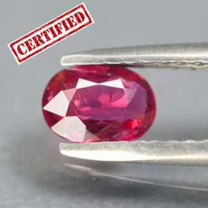 Puede incluir: Una gema de rubí ovalada y facetada sujeta con pinzas. El rubí es de un color rojo rosado intenso con facetas internas visibles. Un sello rojo que dice "CERTIFIED" está en la esquina superior izquierda.