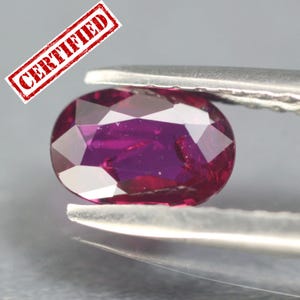 May include: A faceted, oval-shaped, deep red gemstone held by tweezers. The gem has a vibrant, polished surface with multiple facets reflecting light. A red stamp with the word "CERTIFIED" is in the upper left corner.
