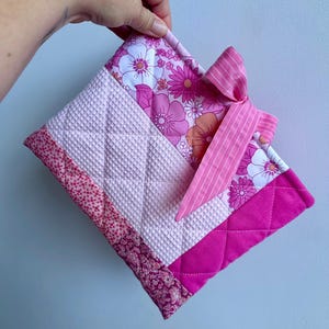 May include: A quilted hot pad in shades of pink, featuring a floral pattern, waffle texture, and a solid pink border. A striped pink bow is attached to the top. The hot pad is held by a hand.
