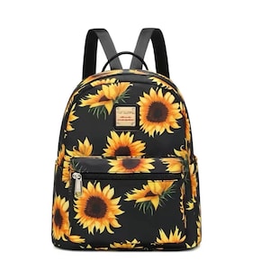 May include: A black backpack featuring a vibrant sunflower pattern with yellow petals and dark brown centers. The bag includes a front zippered pocket, a top handle, and two black shoulder straps. A small rectangular gold-toned metal logo plate is centered.