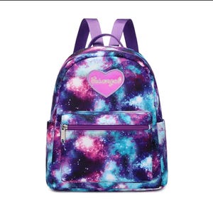 May include: A backpack featuring a vibrant galaxy print in shades of purple, blue, and pink. The bag includes a front zippered pocket and a pink heart-shaped patch on the front with the text "Busangel" written in a cursive white font.