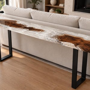 May include: A long, narrow console table featuring a live-edge wood slab integrated with a translucent white resin river design. The tabletop is supported by two rectangular black metal legs, providing a stable base for this furniture piece.