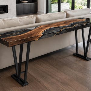 May include: A long console table featuring a live edge wood slab centerpiece with natural brown grain, flanked by black resin sections with gold-colored veining. The table is supported by two black metal V-shaped legs with rectangular base plates.